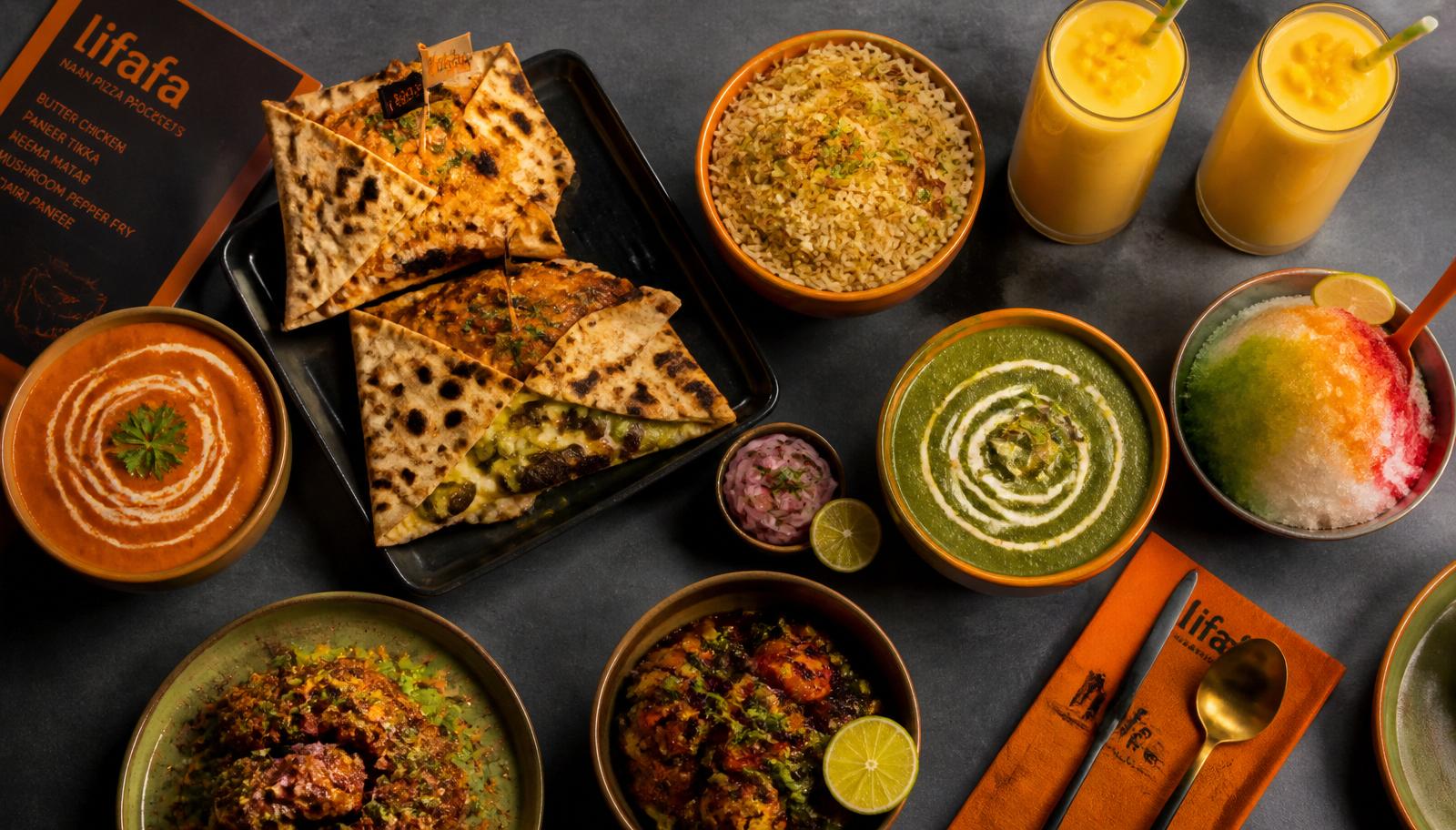 Overhead spread of Indian fusion dishes at FOMO Tampa — Lifafa pizza pockets, bowls, wraps and a mango smoothie on a charcoal table